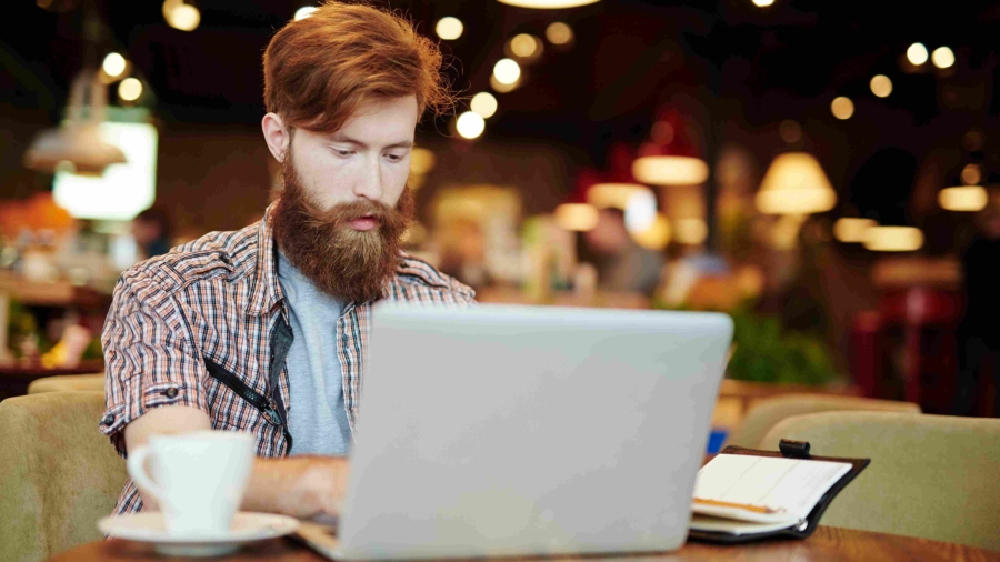 homem branco, de barba e cabelos ruivos usando laptop em um cyber café